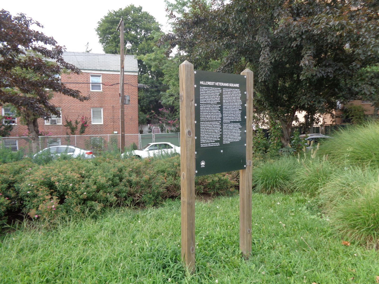 Hillcrest Veterans Square sign amid grasses, shrubs and trees beside Union Turnpike.