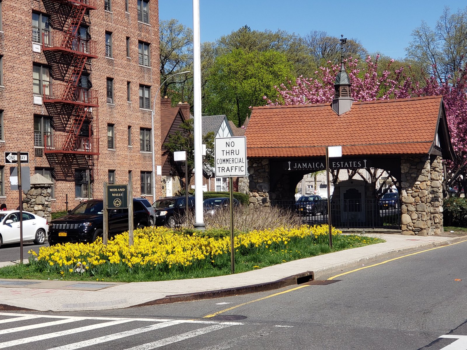 Jamaica Estates gatehouse with stone piers, tiled roof and memorial enclosure beside spring flowers, April 2019.