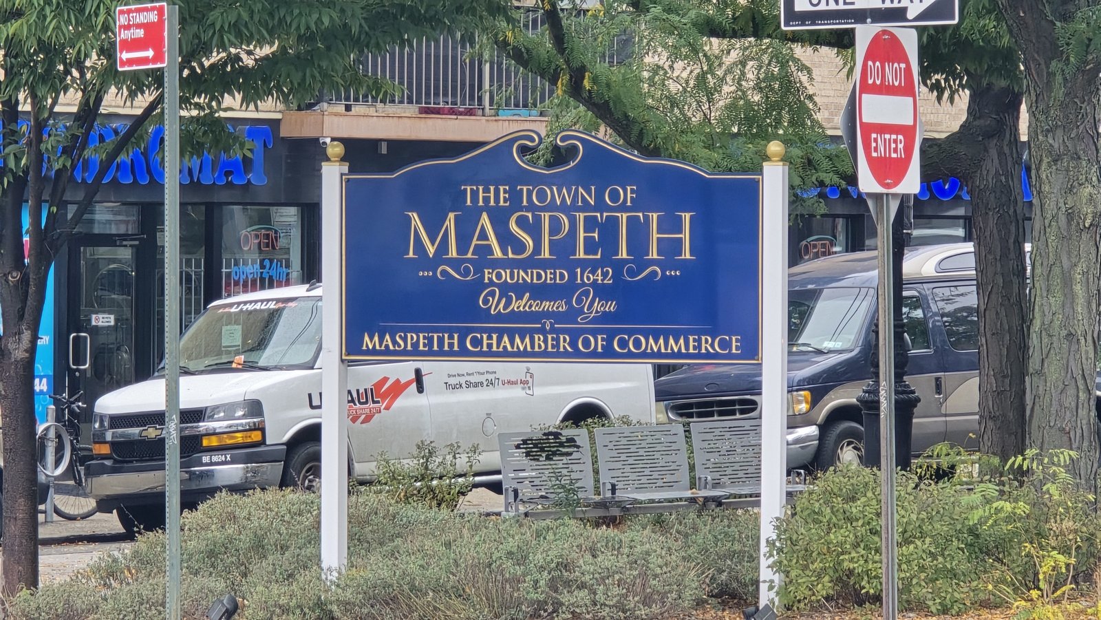 A blue-and-gold Maspeth Chamber of Commerce welcome sign beside a planted street corner.