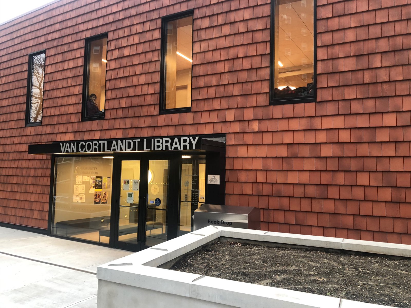 Van Cortlandt Library entrance and sign beneath tall windows in a terracotta-clad facade, photographed in 2020.