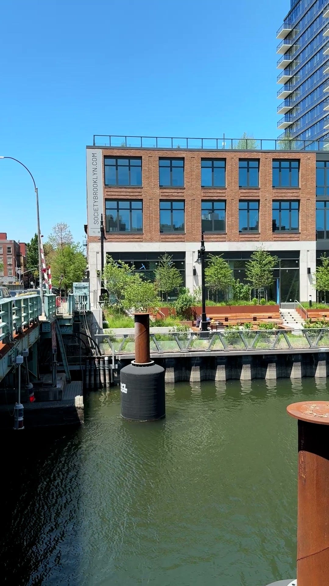 Gowanus Canal water and bridge machinery lie below a planted promenade and brick building with broad windows.