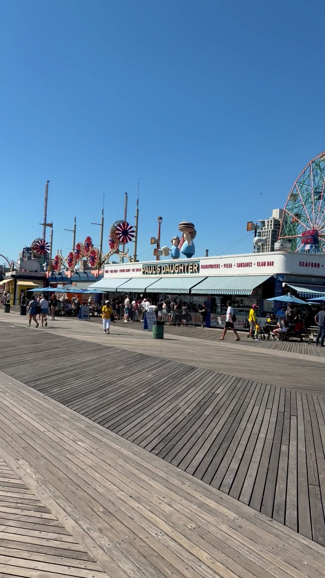 A broad wooden boardwalk passes food stands and colorful amusement rides beneath a clear blue sky.