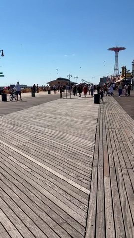 People walk along a wide wooden boardwalk, with a tall red amusement tower rising in the distance.