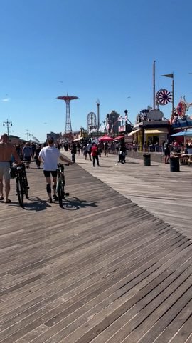 People walk and wheel bicycles along a wooden boardwalk beside amusement rides and food stands.