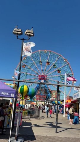 A colorful Ferris wheel rises behind an entrance walkway, flags, balloons and people.