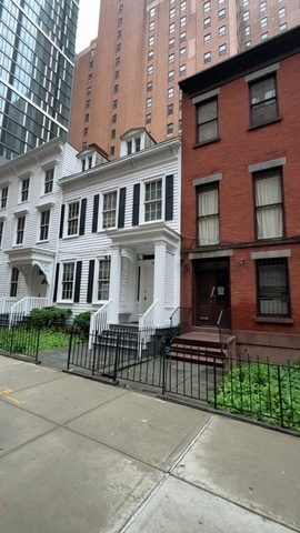 White clapboard houses with porches stand beside a red-brick house, with taller buildings behind them.