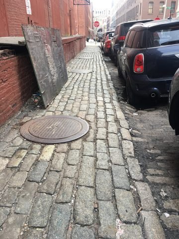 A narrow Belgian-block sidewalk runs beside a brick wall and parked cars.