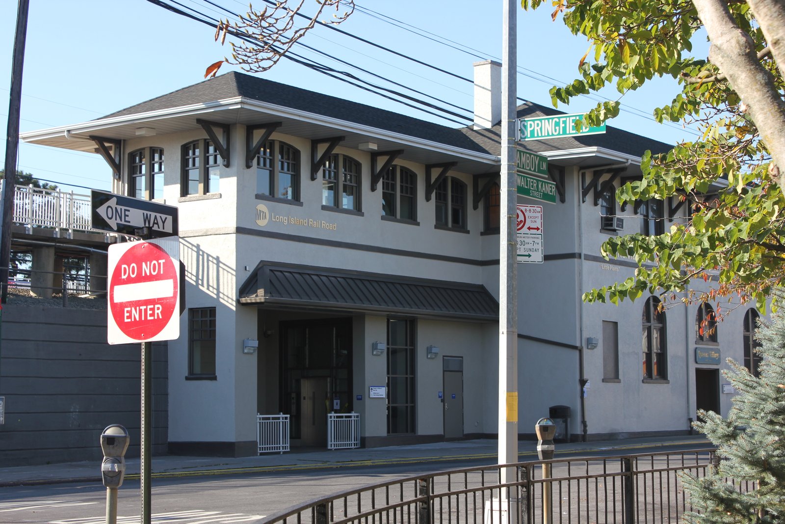 Queens Village station building beside Springfield Boulevard and Amboy Lane, with cream walls, dark roof and arched windows.
